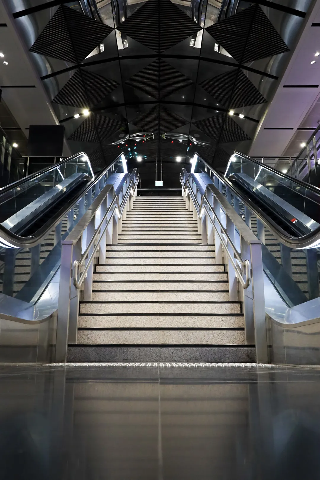 A symmetrical pair of escalators and stairs beneath a mirrored ceiling