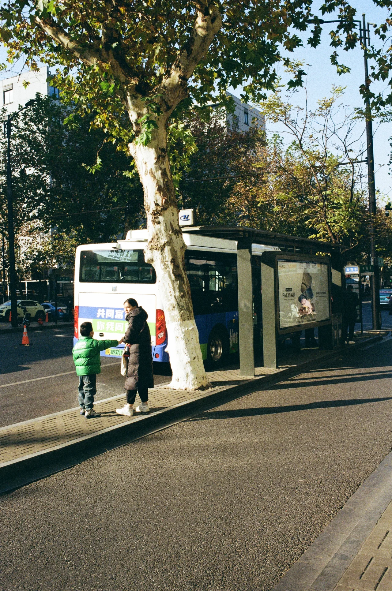 An adult and child waiting beside a city bus stop in late-afternoon light
